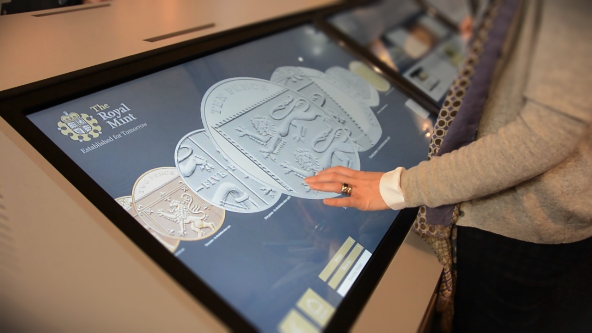 woman interacting with The Royal Mint interactive touchscreen software displayed on large monitor on white surface