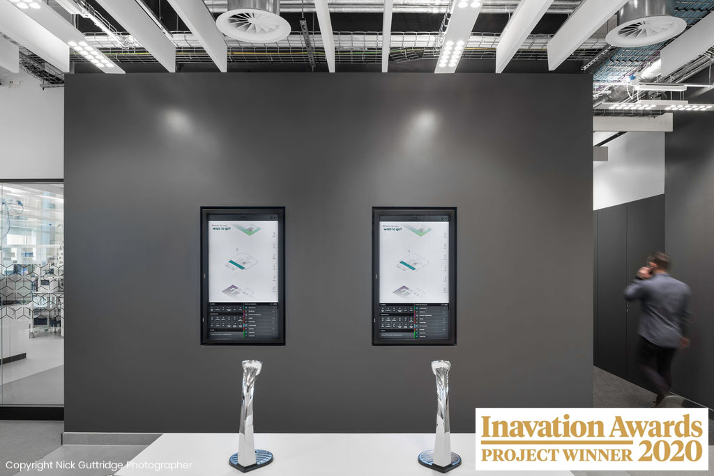 Two interactive touchscreens displayed on a dark grey walled office space with two Inavation Awards for project winner 2020 in front